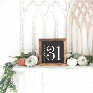 May include: A wooden framed chalkboard with the number 31 in white, with a spider and floral accents. The chalkboard is surrounded by pumpkins and greenery, creating a festive Halloween display.
