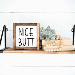 May include: A wooden shelf with black metal brackets holds a framed sign that reads "NICE BUTT" in black script. The shelf is decorated with a small potted plant and a strand of wooden beads.