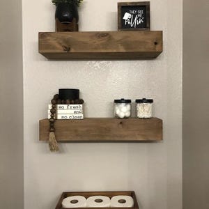 May include: Two wooden shelves with a black sign that says "They see me rollin'" on the top shelf. The bottom shelf has a stack of small white blocks with the words "so fresh and so clean" on them. There are two clear glass jars with black lids on the bottom shelf, one with cotton balls and the other with cotton swabs.