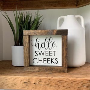 Peut inclure: Une pancarte en bois avec le texte "hello sweet cheeks" en lettres noires sur fond blanc. La pancarte est pos&eacute;e sur une &eacute;tag&egrave;re en bois.