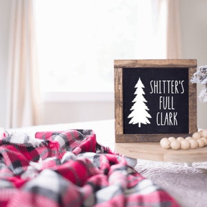 May include: A black framed sign with white text that reads "Shitter's Full Clark" and a white silhouette of a Christmas tree. The sign is sitting on a wooden tiered tray with a small, snowy artificial Christmas tree in a brown pot.