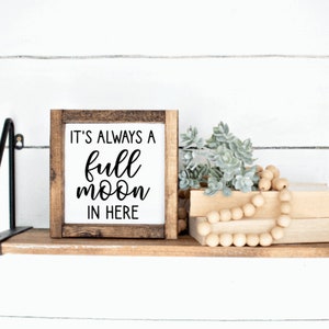 May include: A wooden sign with the text "It's always a full moon in here" hanging on a shelf with a small plant and wooden beads.