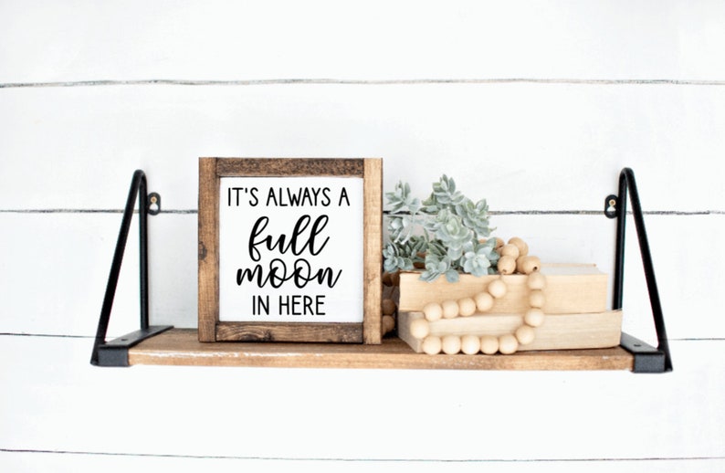 May include: A wooden sign with the text "It's always a full moon in here" hanging on a shelf with a small plant and wooden beads.