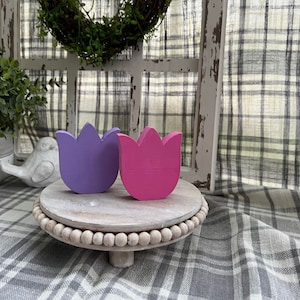 May include: Two wooden tulip-shaped decorations, one purple and one pink, are displayed on a white wooden tiered stand with a beaded edge.