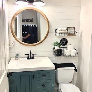 Peut inclure: Une vasque de salle de bain blanche avec un meuble bleu et un plan de travail blanc. La vasque est surmont&eacute;e d'un miroir rond avec un cadre en bois. Deux &eacute;tag&egrave;res en m&eacute;tal noir sont plac&eacute;es au-dessus de la vasque, avec des serviettes blanches, du papier toilette et une petite plante. Les &eacute;tag&egrave;res portent des pancartes qui disent "hello sweet cheeks" et "wash your hands".