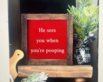 以下が含まれることがあります： 赤い背景に白い文字で「He sees you when you're pooping」と書かれた素朴な木製の看板。看板は濃い色の木枠で囲まれ、旋盤で削られた脚のある木製の棚の上に置かれている。