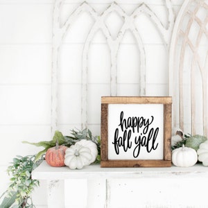 May include: A wooden sign with the text "happy fall y'all" in black script. The sign is sitting on a white shelf with a green garland and white pumpkins.