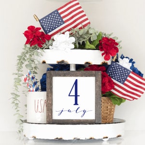 May include: A tiered tray decorated with red, white and blue for the Fourth of July. The tray features a sign that says "4 July" in blue lettering, red and blue striped straws, a white mug with the word "USA" in red lettering, and small American flags.