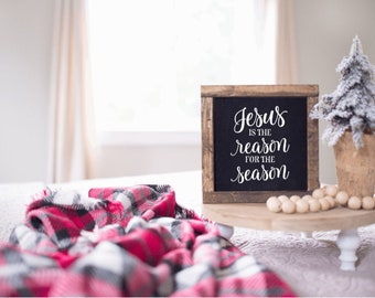 Puede incluir: Un letrero de madera con el texto "Jesús es la razón de la temporada" en letras blancas sobre fondo negro. El letrero está sentado en una bandeja de madera con un pequeño árbol de Navidad cubierto de nieve en una maceta y cuentas de madera.