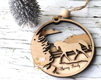 Puede incluir: Adorno de caballo de madera con silueta de caballo, árboles y la inscripción "Murray Family". El adorno redondo tiene un acabado de madera natural y una cuerda de yute para colgar. Un pequeño árbol de Navidad plateado está en el fondo.