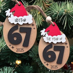 May include: Two wooden Christmas tree decorations with a Santa hat design. One says "Hector" and the other says "The Year of". Both feature the number "67" in black, the year "2025", and star accents. They hang from a twine string.