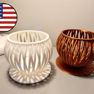 May include: Two decorative candle holders. One is white, the other is copper-colored. Both have a spherical shape with a lattice design. The white candle holder is lit from within, casting a patterned shadow on the surface below.