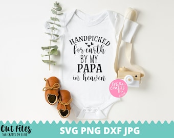 May include: White baby onesie with black text that reads "Handpicked for earth by my Papa in heaven". The onesie is on a white background with a small wooden toy camera and two brown baby shoes.