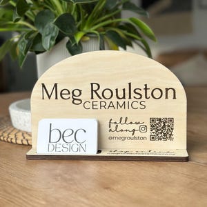 Peut inclure: Un présentoir en bois clair en forme de demi-cercle avec le texte gravé au laser Meg Roulston CERAMICS, un code QR et le compte @megroulston. Une carte de visite blanche avec le texte bec DESIGN est insérée dans la base avant du support.