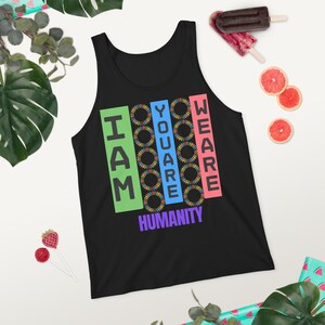 Può includere: Canotta nera con la scritta "I AM YOU ARE WE ARE HUMANITY" in blocchi verticali colorati. Il design include grafiche a forma di girasole. La canotta è esposta su una superficie bianca con elementi decorativi.
