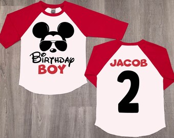 May include: A white and red baseball-style shirt with a black and white Mickey Mouse wearing sunglasses on the front. The text "2nd Birthday Boy" is printed in black and red. The back of the shirt has the name "Jacob" and the number "2" printed in red.