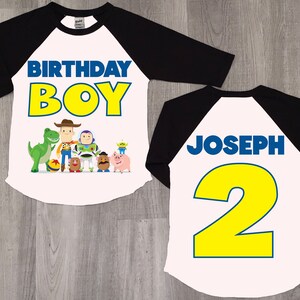 K&ouml;nnte beinhalten: Ein wei&szlig;es und schwarzes Baseball-Shirt mit Toy Story-Motiv. Die Vorderseite des Shirts tr&auml;gt die Aufschrift "Birthday Boy" in gelben Buchstaben mit blauem Rand. Die R&uuml;ckseite des Shirts tr&auml;gt die Aufschrift "Joseph 2" in blauen Buchstaben mit gelbem Rand.