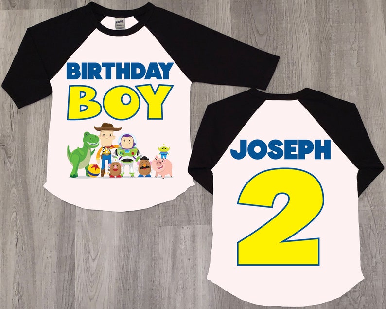 K&ouml;nnte beinhalten: Ein wei&szlig;es und schwarzes Baseball-Shirt mit Toy Story-Motiv. Die Vorderseite des Shirts tr&auml;gt die Aufschrift "Birthday Boy" in gelben Buchstaben mit blauem Rand. Die R&uuml;ckseite des Shirts tr&auml;gt die Aufschrift "Joseph 2" in blauen Buchstaben mit gelbem Rand.