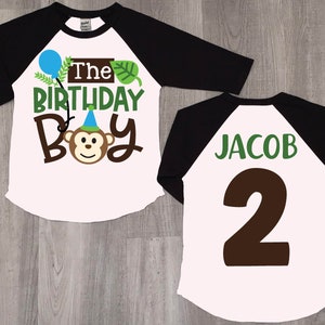 May include: White and black baseball style shirt with a monkey graphic and the text "The Birthday Boy". The back of the shirt has the name "Jacob" and the number "2" in brown.