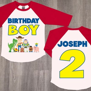 K&ouml;nnte beinhalten: Rotes und wei&szlig;es Baseball-Shirt mit Toy Story-Motiv. Die Vorderseite des Shirts tr&auml;gt den Schriftzug "Birthday Boy" mit einer Gruppe von Toy Story-Charakteren. Die R&uuml;ckseite des Shirts tr&auml;gt den Schriftzug "Joseph 2" in Blau und Gelb.