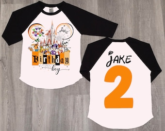 Puede incluir: Camiseta de estilo béisbol blanca y negra con un gráfico de personajes de Disney con temática de Halloween en la parte delantera. El gráfico incluye a Mickey Mouse, Minnie Mouse, Goofy, Pluto, Donald Duck y otros personajes de Disney. El texto "Birthday Boy" está impreso debajo del gráfico. La parte posterior de la camiseta tiene el nombre "Jake" y el número "2" impreso en naranja.