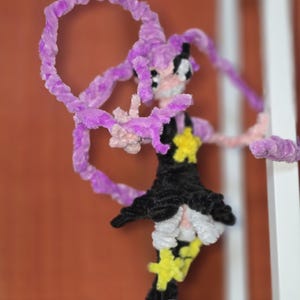 May include: A handmade pipe cleaner doll with a black dress, yellow star accents, and purple hair and accents. The doll has a whimsical design and is posed with arms outstretched. The background is a mix of white and orange.
