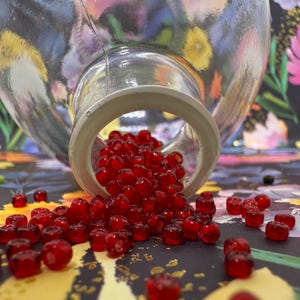 May include: A clear glass jar tipped over, spilling out numerous small, red, bead-like objects. The jar's opening is visible, with the beads scattered on a floral patterned surface. The beads are a deep, translucent red.