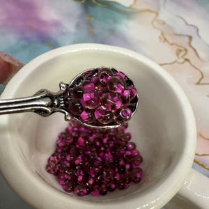 Puede incluir: Un cuenco de cerámica blanco con asa está lleno de pequeñas cuentas redondas, rosas y transparentes. Una cuchara decorativa plateada contiene una cucharada de cuentas, con más cuentas que se vierten en el cuenco. El fondo tiene un diseño veteado.