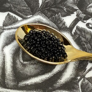 May include: A gold-colored spoon filled with small, black beads. The spoon rests on a black and white floral patterned background. The beads appear to be seed beads, often used in jewelry making and crafts.