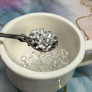 Puede incluir: Una taza de cerámica blanca llena de cuentas redondas y transparentes. Una cuchara decorativa de plata contiene una cucharada de cuentas, colocada sobre la taza. El fondo presenta un diseño abstracto colorido con detalles dorados.