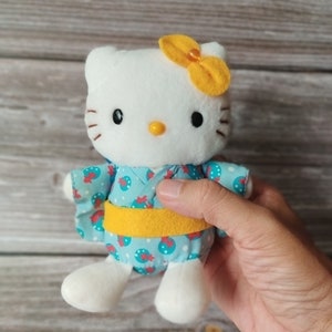 Pode incluir: Um gato de pelúcia branco com um laço amarelo, vestindo um quimono azul com um padrão vermelho e branco. O quimono tem um cinto amarelo. O gato tem olhos pretos, bigodes e um nariz amarelo.
