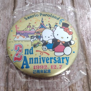 May include: A round, gold-colored pin commemorating the 2nd Anniversary of Sanrio Puroland. The pin features cartoon characters, the text "2nd Anniversary 1992.12.7", and the Sanrio Puroland logo. The pin is sealed in a clear plastic wrapper.