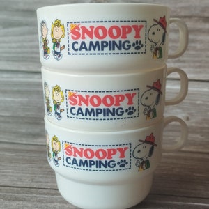 May include: Three white ceramic mugs stacked on top of each other. Each mug features the words "SNOOPY CAMPING" in red and blue, along with cartoon characters. The mugs have a handle and a rounded bowl shape.
