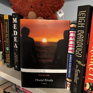 Pode incluir: Uma estante com vários livros, incluindo "Heated Rivalry" de Rachel Reid, publicado pela Penguin Classics. A capa apresenta silhuetas contra um pôr do sol. Um brinquedo de folha de pelúcia é visível.