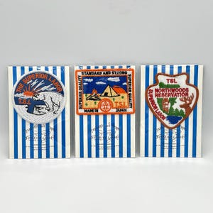 May include: Three embroidered patches on blue and white striped backing cards. The left patch is circular with a white bear and sun. The center patch is rectangular with a tent and campfire. The right patch is shield-shaped with a deer and forest.