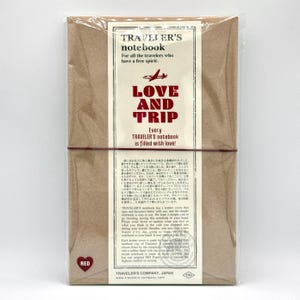 May include: A brown Traveler's Notebook, wrapped in clear plastic, with a red elastic band. The label reads "TRAVELER'S notebook" and "LOVE AND TRIP." The notebook is designed for travelers and has a leather cover.