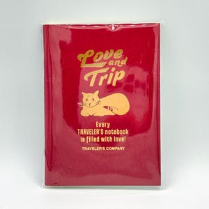 May include: A red notebook with the words "Love and Trip" in gold lettering, along with a gold cat illustration. The text "Every Traveler's notebook is filled with love!" is also present. The notebook is in a clear plastic sleeve.