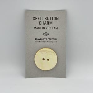 May include: A white shell button charm with two holes. The charm is on a card that says "Shell Button Charm" and "Made in Vietnam". The card also has the logo "TFA" for Traveler's Factory and the website address www.travelers-factory.com.