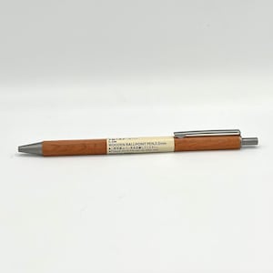 May include: A wooden ballpoint pen with a silver clip and a brown wooden barrel. The pen has a 0.5mm tip and the text "WOODEN BALLPOINT PEN, 0.5mm" is printed on the barrel. The text "ご使用後はペン先を収納してください。" is also printed on the barrel.
