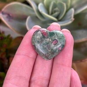 May include: A heart-shaped stone, predominantly green with red flecks. The stone is held in a hand, with a blurred background of green succulent plants.