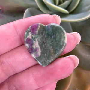 May include: A heart-shaped gemstone with a green base and patches of red. The stone is held in a hand, with a succulent plant in the background. The gemstone is approximately 4 cm wide.