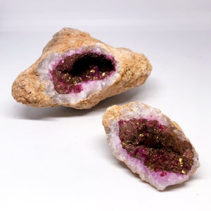 May include: Two geode halves with a pink and gold interior. The geode halves are sitting on a white surface.