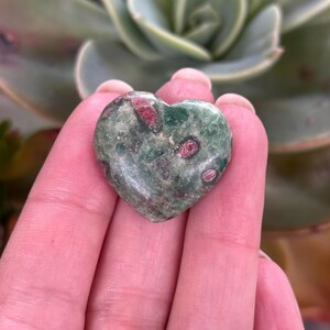May include: A heart-shaped stone with a green base and red inclusions. The stone is held in a hand, with a succulent plant in the background. The stone appears polished and smooth.