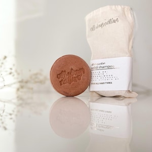 May include: A round, brown solid shampoo bar with the text "allways nature" embossed on it. The bar is sitting on a white surface next to a white cloth bag with the text "allways nature" printed on it.