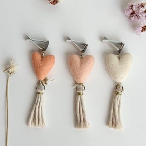 May include: Three heart-shaped car air fresheners in peach, pink, and cream colors. Each has a silver clip, gold ring, and a cream-colored tassel. The hearts are made of felt. Dried flowers and a white background complete the image.
