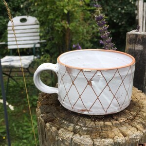 Könnte beinhalten: Eine handgefertigte weiße Keramik-Tasse mit Rautenmuster und braunem Rand. Die Tasse hat einen gebogenen Henkel und steht auf einem verwitterten Holzstumpf. Der Hintergrund umfasst Grün und einen weißen Stuhl.
