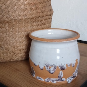Könnte beinhalten: Weißer Keramiktopf mit braunem Rand und Boden. Der Boden hat ein strukturiertes, abstraktes Design in Braun- und Lilatönen. Der Topf steht auf einer Holzoberfläche neben einem geflochtenen Korb.