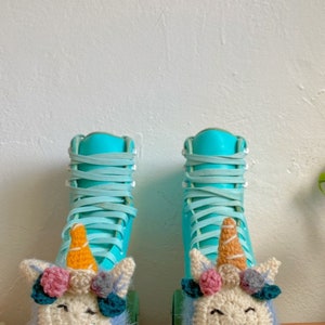 May include: A pair of teal roller skates with crocheted unicorn toppers. The unicorns have pink and blue flowers, brown horns, and fuzzy white fur.