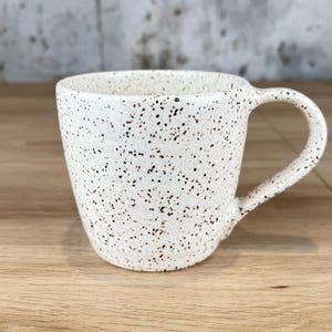 May include: A speckled white ceramic mug with a rounded square shape and a curved handle. The mug is covered in small, dark brown speckles. The mug is sitting on a wooden surface.