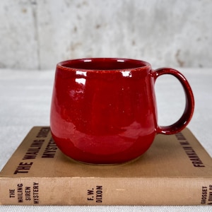 May include: A glossy, red ceramic mug with a rounded body and a handle. The mug sits atop a vintage book titled "The Wailing Siren Mystery" by F.W. Dixon. The mug's interior is also red.